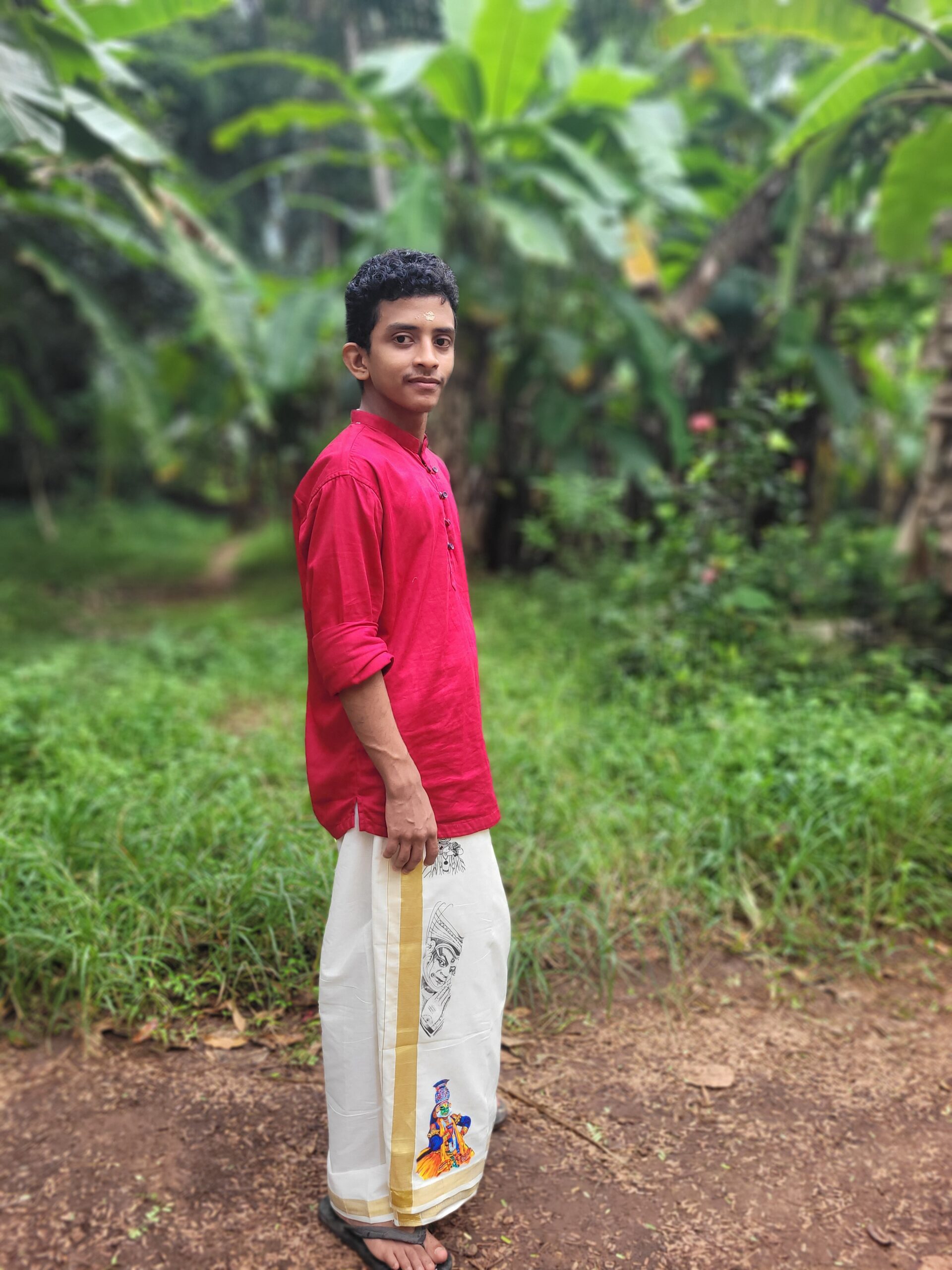 Portrait of a young Indian man wearing a traditional red Kurta and a white, decorated Kerala style Mundu (dhoti) outdoors near banana trees and grass.